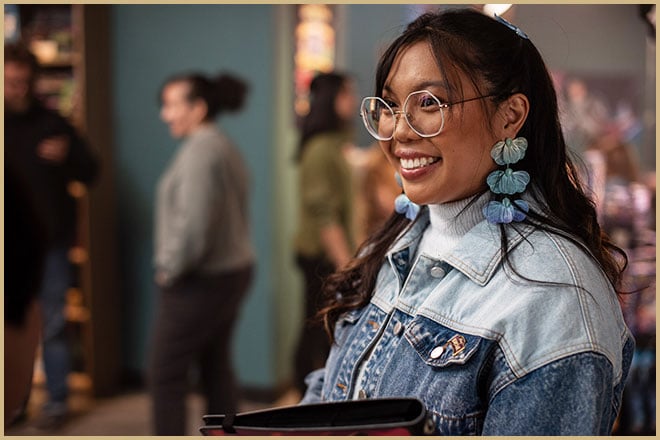 Photo of a young woman smiling and standing in a game store while she carried her Disney Lorcana Trading Card Game card portfolio